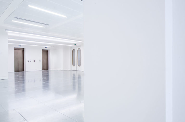 An interior of an open office space painted white with silver sub flooring. theres 2 lifts on one wall and a door on another. London Interior photographer | David Chatfield