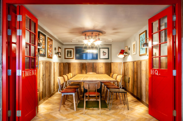 Restaurant interior called the Hydrant in London. behind red doors theres a big table with seats of many styles around it. Light in the corner and a cluster of lights on the ceiling. interior photographer | david chatield photography