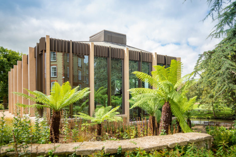 Architectural image of building called St Hildas, Oxford university. made of glass, steel and stone, planted heavily with dicksonias and other tropical shrubs. Blue sky with white clouds as a background. architectural photographer, David Chatfield