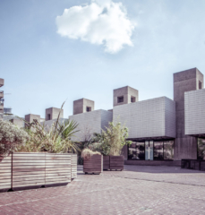 Brutalist building with geometric forms and textured facade, surrounded by large planters on a paved courtyard. Cloud and blue sky in the background.