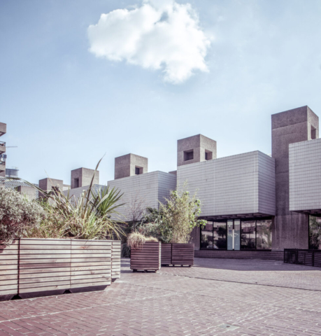 Brutalist building with geometric forms and textured facade, surrounded by large planters on a paved courtyard. Cloud and blue sky in the background.