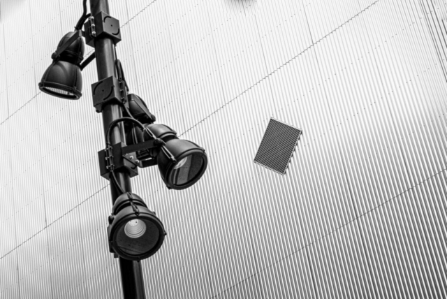 Black and white image of a pole with multiple spotlights against a textured, corrugated metal wall with a small rectangular vent.