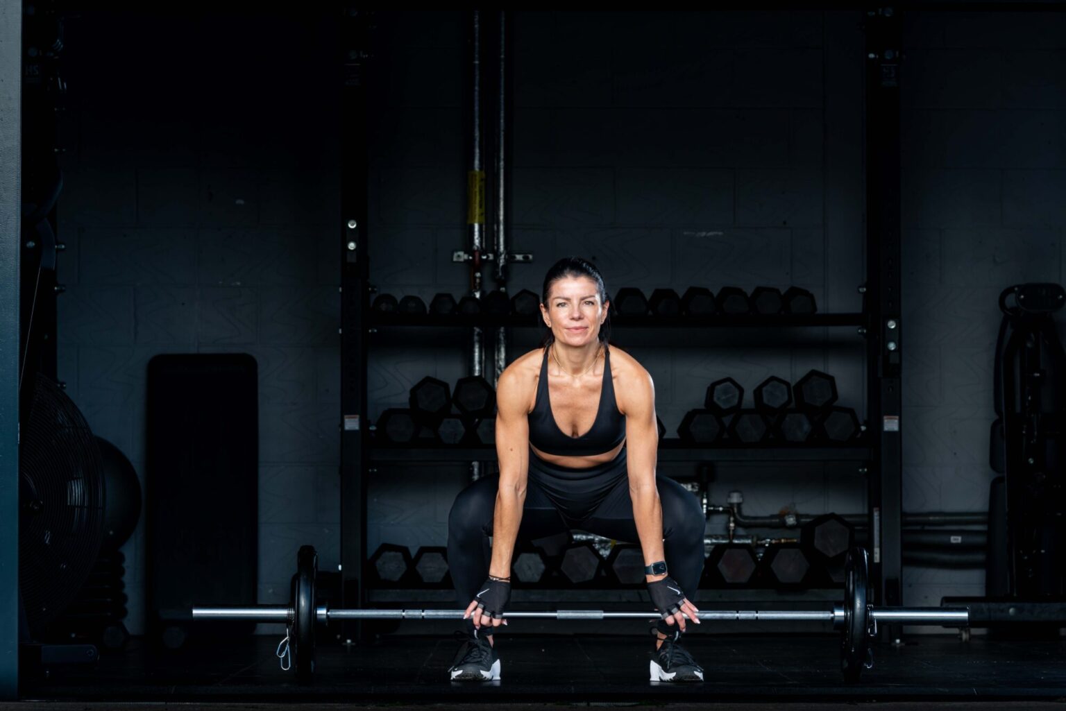 female in a gym setting working out with a barbell, weaing black lycra leggings and black top low key background. weights behind the model
