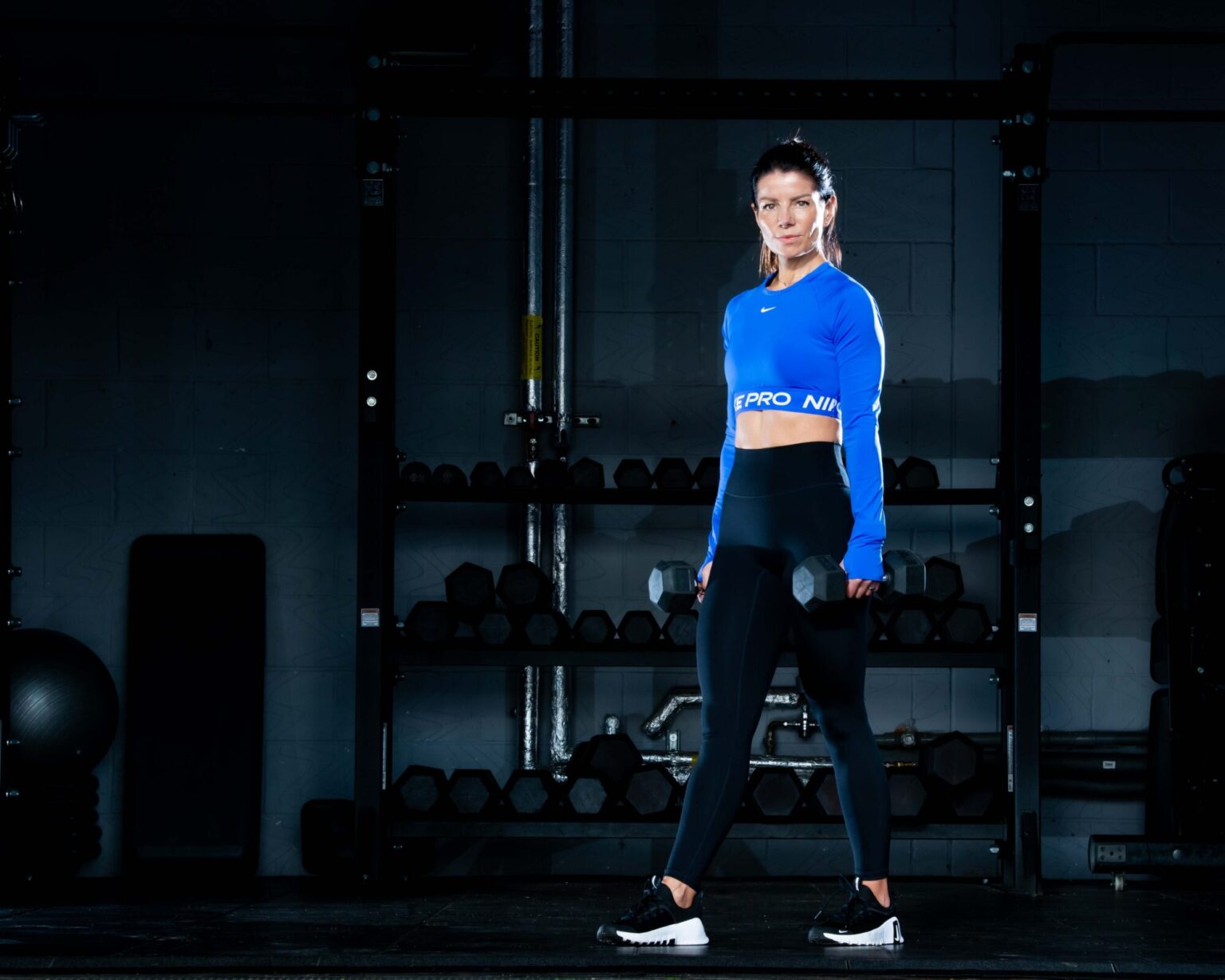 female in a gym setting working out with dumbbells, weaing black lycra leggings and blue top, low key background.