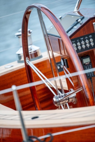 The wheel of a mahogany yacht. the wheel has a chrome center and wooden outer wheel. water in the background.