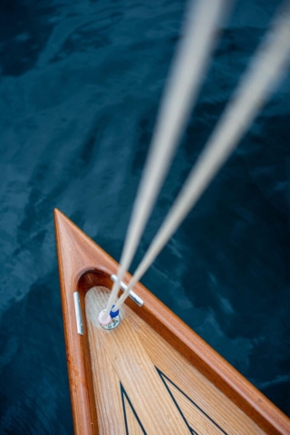 looking down on the front of a small tinmber yacht. to ropes leading from top right of the screen to the middle attaching on the front of the boat. the water is dark blue and very calm.